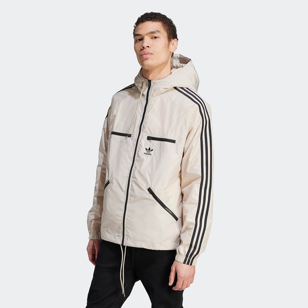 Herren-Model trägt eine khaki adidas Originals Windbreaker Jacke mit schwarzen 3-Streifen und Trefoil-Logo vor dem STICKABUSH Store in Berlin-Kreuzberg.