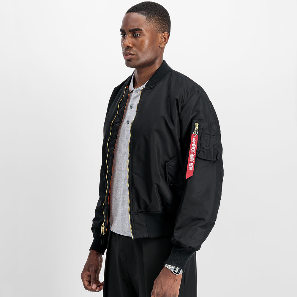 “Alpha Industries MA-1 (HERITAGE) Bomberjacke in Schwarz, aus robustem Nylon, mit ikonischem Flight-Tag am Ärmel und gestepptem orangenem Innenfutter.“