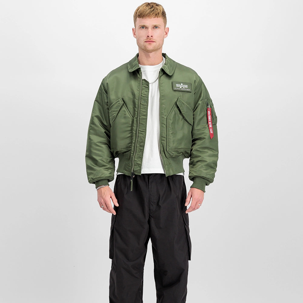 „Alpha Industries CWU 45 Heritage Bomberjacke in Sage Green mit Umlegekragen, Klettverschluss-Taschen und rotem Remove Before Flight Tag bei Stickabush Berlin.“