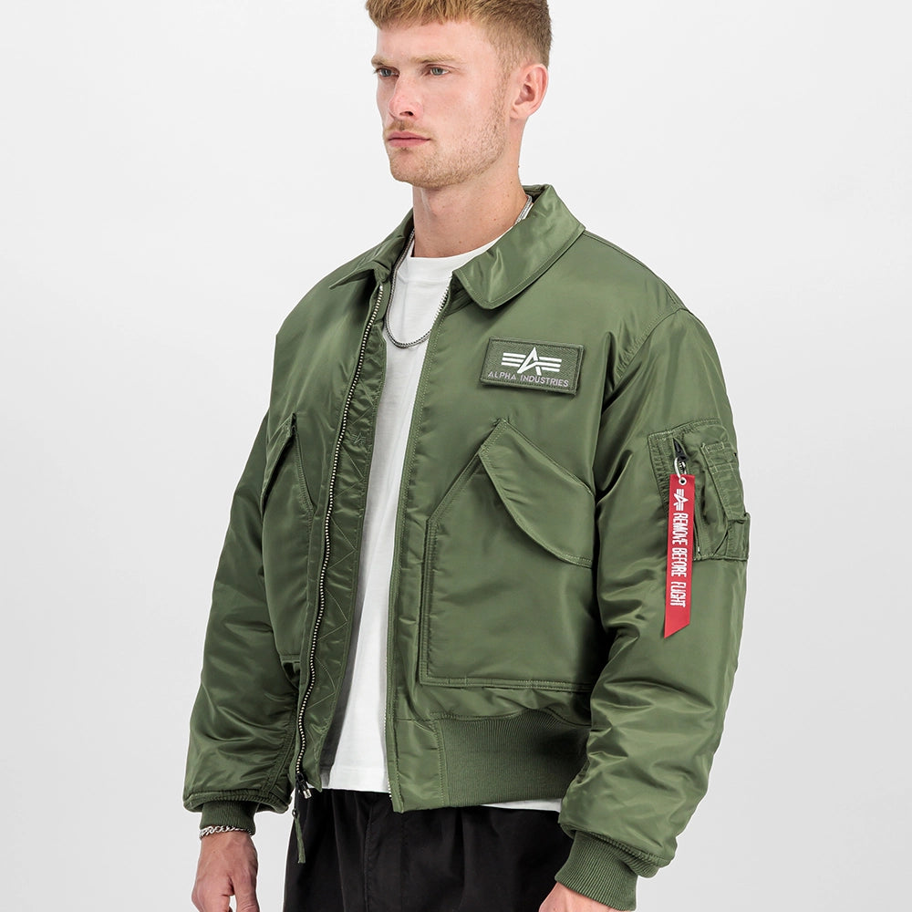 „Alpha Industries CWU 45 Heritage Bomberjacke in Sage Green mit Umlegekragen, Klettverschluss-Taschen und rotem Remove Before Flight Tag bei Stickabush Berlin.“
