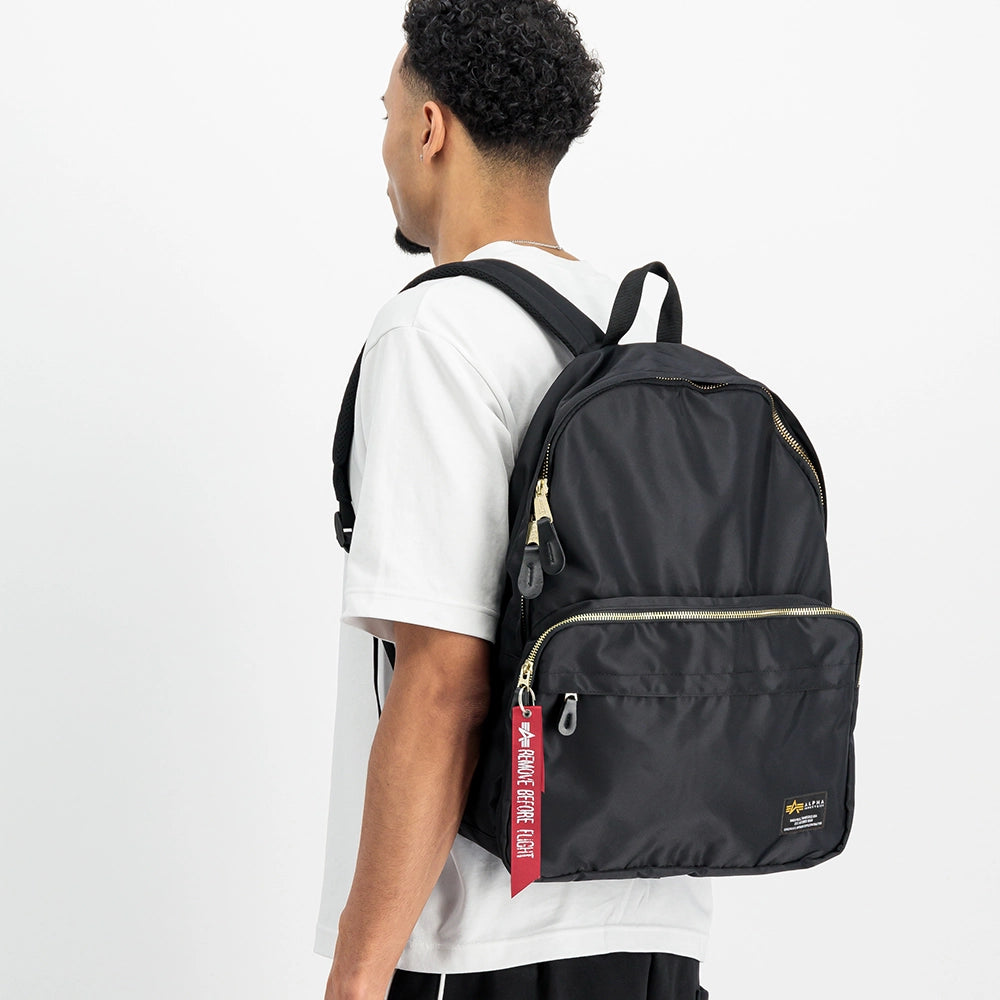 “Schwarzer Alpha Industries Crew Backpack aus strapazierfähigem Flight-Nylon mit rotem Remove Before Flight Anhänger und gepolsterten Trägern bei Stickabush Berlin.“