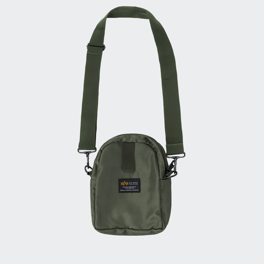 “Grüne (Sage Green) Alpha Industries Crew Carry Bag Umhängetasche mit rotem Remove Before Flight Anhänger und verstellbarem Schultergurt bei Stickabush Berlin.“