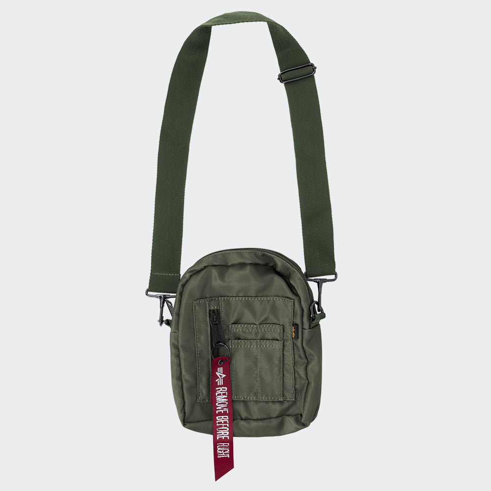 “Grüne (Sage Green) Alpha Industries Crew Carry Bag Umhängetasche mit rotem Remove Before Flight Anhänger und verstellbarem Schultergurt bei Stickabush Berlin.“