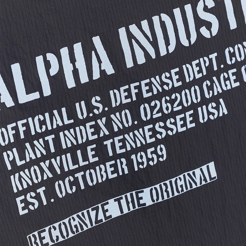 “Schwarzes Alpha Industries Packable Rain Cape mit Logo auf der Brust, verstellbarer Kapuze und rotem Remove Before Flight Anhänger bei Stickabush Berlin.“