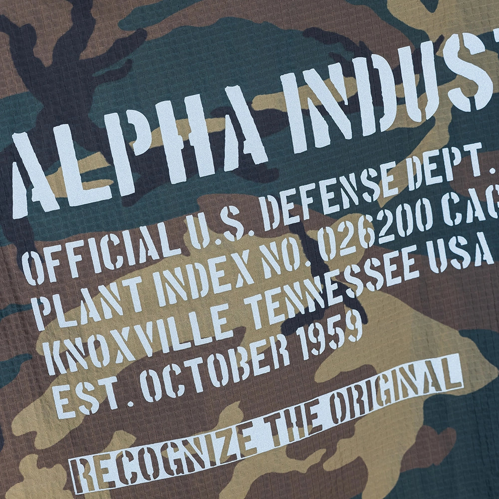 “Alpha Industries Regenponcho im Woodland Camouflage Muster mit verstellbarer Kapuze und rotem Remove Before Flight Anhänger bei Stickabush Berlin.“