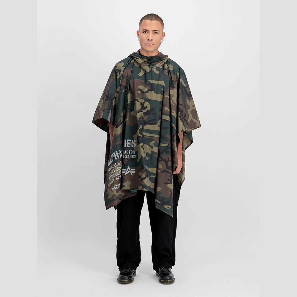 “Alpha Industries Regenponcho im Woodland Camouflage Muster mit verstellbarer Kapuze und rotem Remove Before Flight Anhänger bei Stickabush Berlin.“
