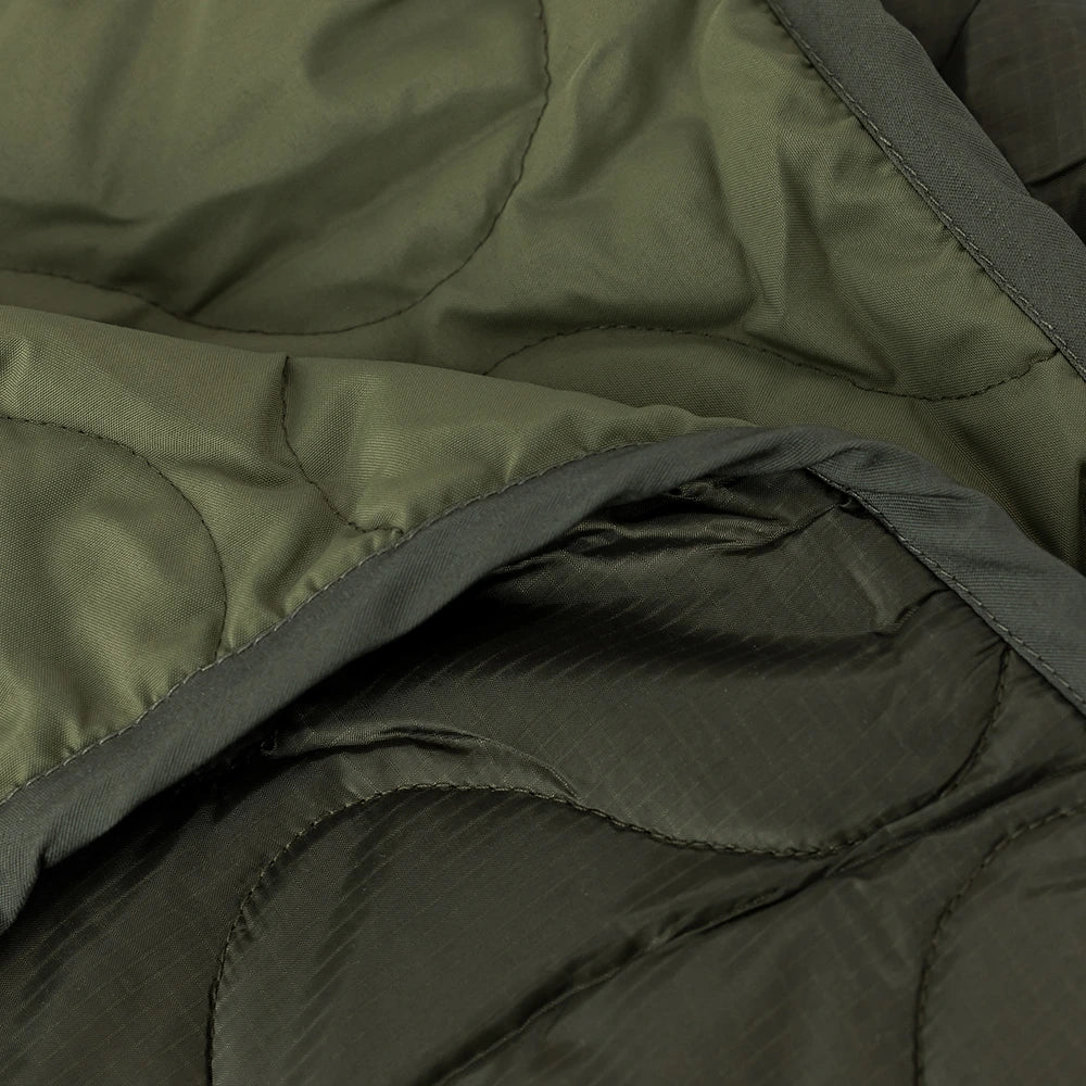 “Dunkelgrüne (Dark Olive) Alpha Industries Picknickdecke aus Flight-Nylon, zusammengefaltet in der integrierten Tasche mit rotem Remove Before Flight Tag bei Stickabush Berlin.“