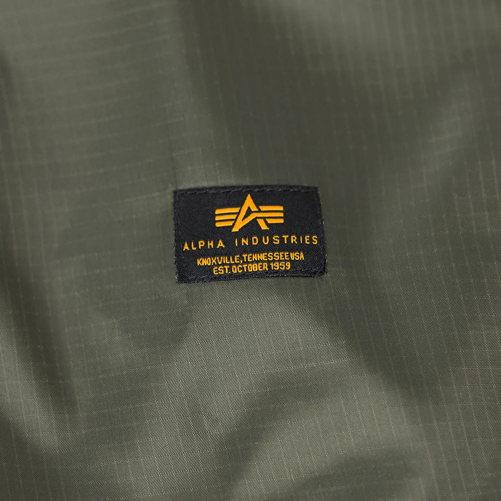 “Dunkelgrüne (Dark Olive) Alpha Industries Picknickdecke aus Flight-Nylon, zusammengefaltet in der integrierten Tasche mit rotem Remove Before Flight Tag bei Stickabush Berlin.“