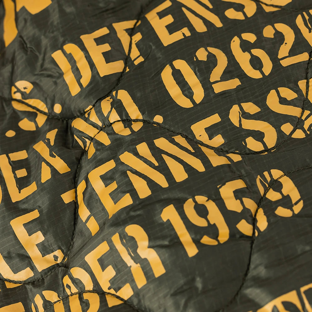 “Dunkelgrüne (Dark Olive) Alpha Industries Picknickdecke aus Flight-Nylon, zusammengefaltet in der integrierten Tasche mit rotem Remove Before Flight Tag bei Stickabush Berlin.“