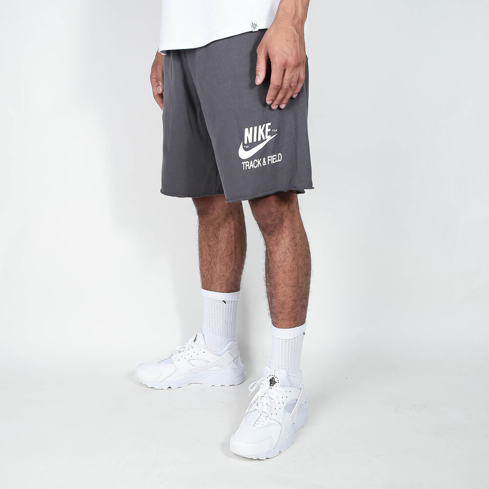 „Nike Sportswear Club Alumni Shorts in Grau/Schwarz mit ungesäumten Kanten, weißem Nike Track & Field Logo-Print und elastischem Bund bei Stickabush Berlin.“