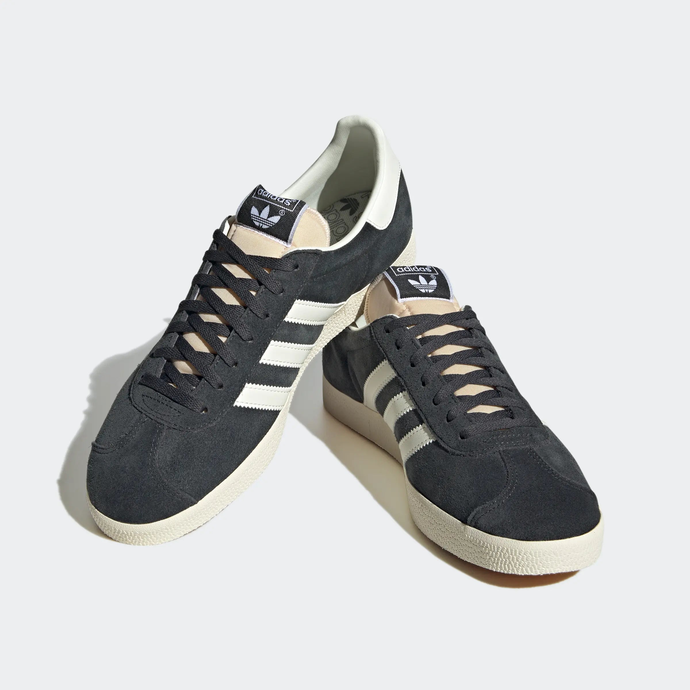 "Anthrazitfarbene Adidas Gazelle Schuhe mit off-weißen Streifen und cremefarbener Sohle. Klassisches Wildleder-Obermaterial, goldener 'Gazelle' Schriftzug an der Seite."