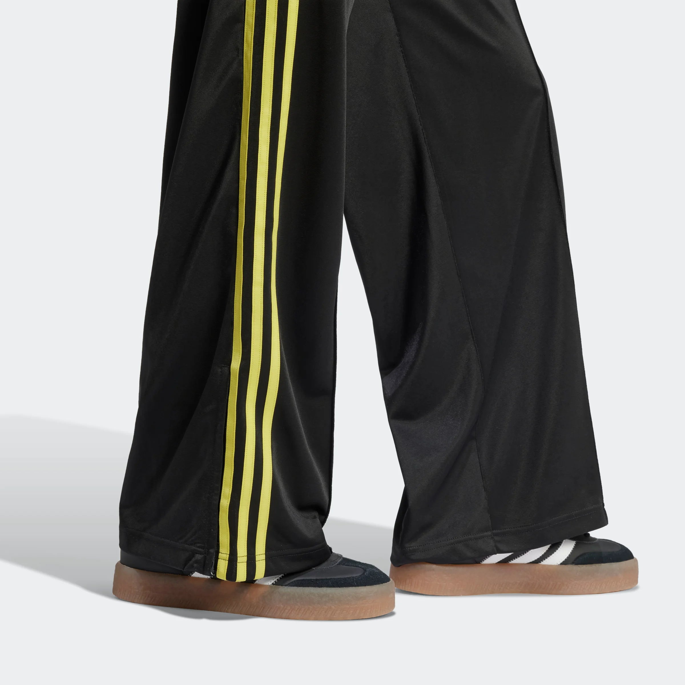 "Schwarze Adidas Firebird Trainingshose für Damen mit hellgelben Seitenstreifen. Lockerer Schnitt, glänzendes Trikotmaterial, seitliche Reißverschlusstaschen, kleines Logo."