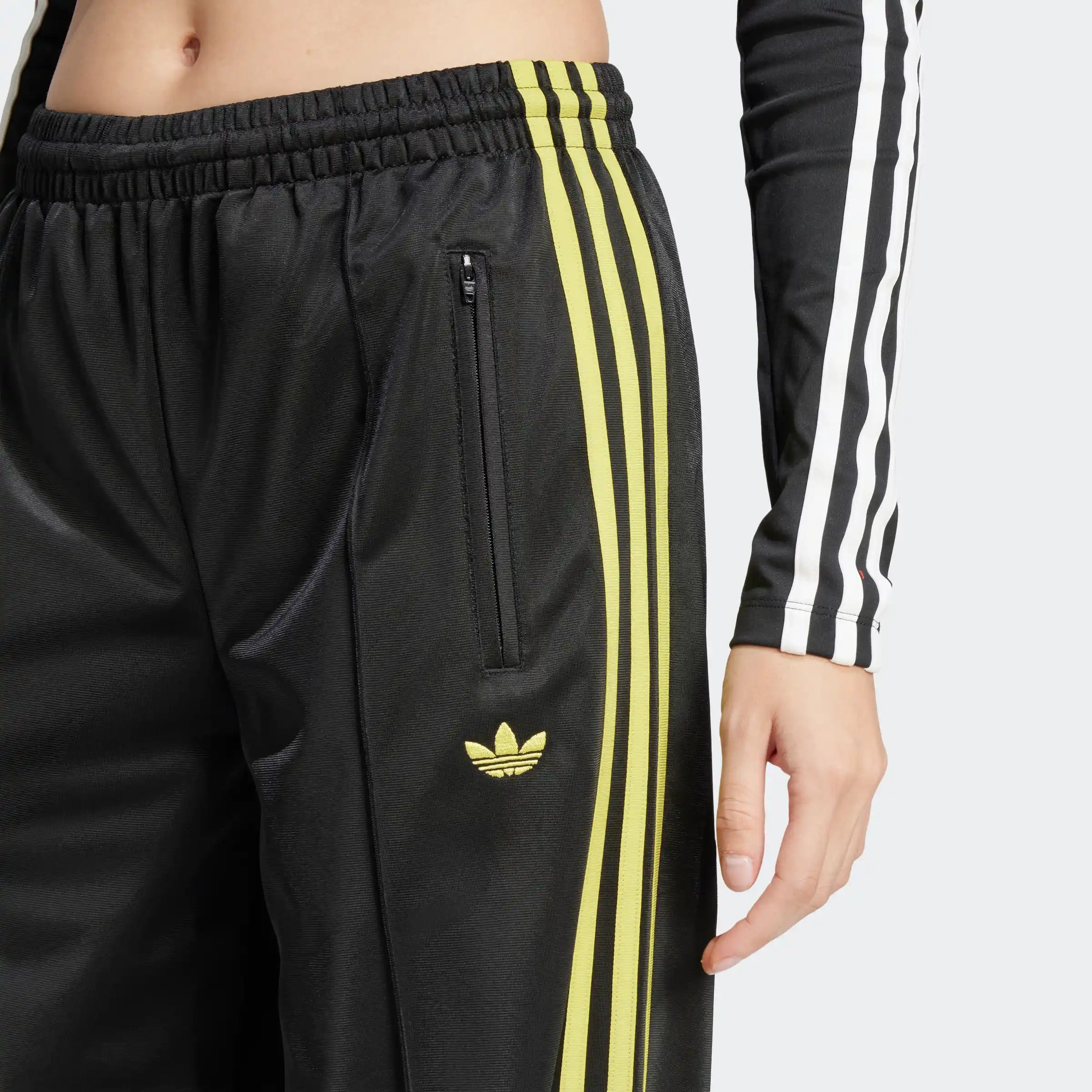 "Schwarze Adidas Firebird Trainingshose für Damen mit hellgelben Seitenstreifen. Lockerer Schnitt, glänzendes Trikotmaterial, seitliche Reißverschlusstaschen, kleines Logo."