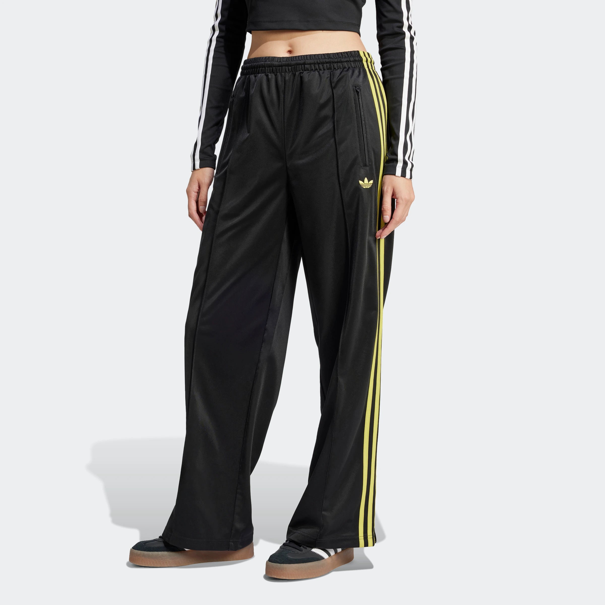 "Schwarze Adidas Firebird Trainingshose für Damen mit hellgelben Seitenstreifen. Lockerer Schnitt, glänzendes Trikotmaterial, seitliche Reißverschlusstaschen, kleines Logo."