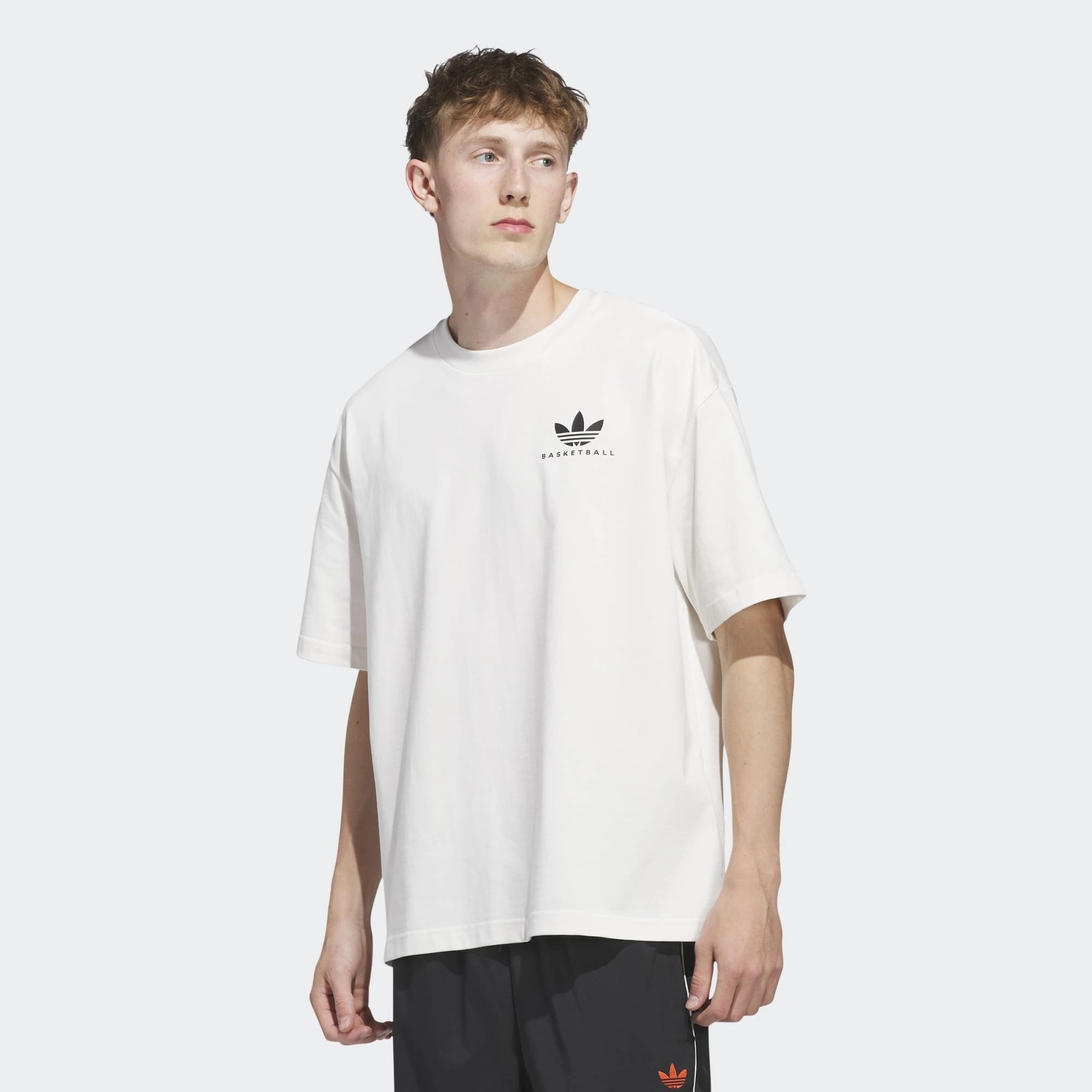 "Vorderansicht am Model des Adidas Originals Worldwide Hoops GFX Tee in Cloud White – Weißes Baumwoll-T-Shirt mit schwarzem Basketball-Grafikprint bei Stickabush Berlin.“