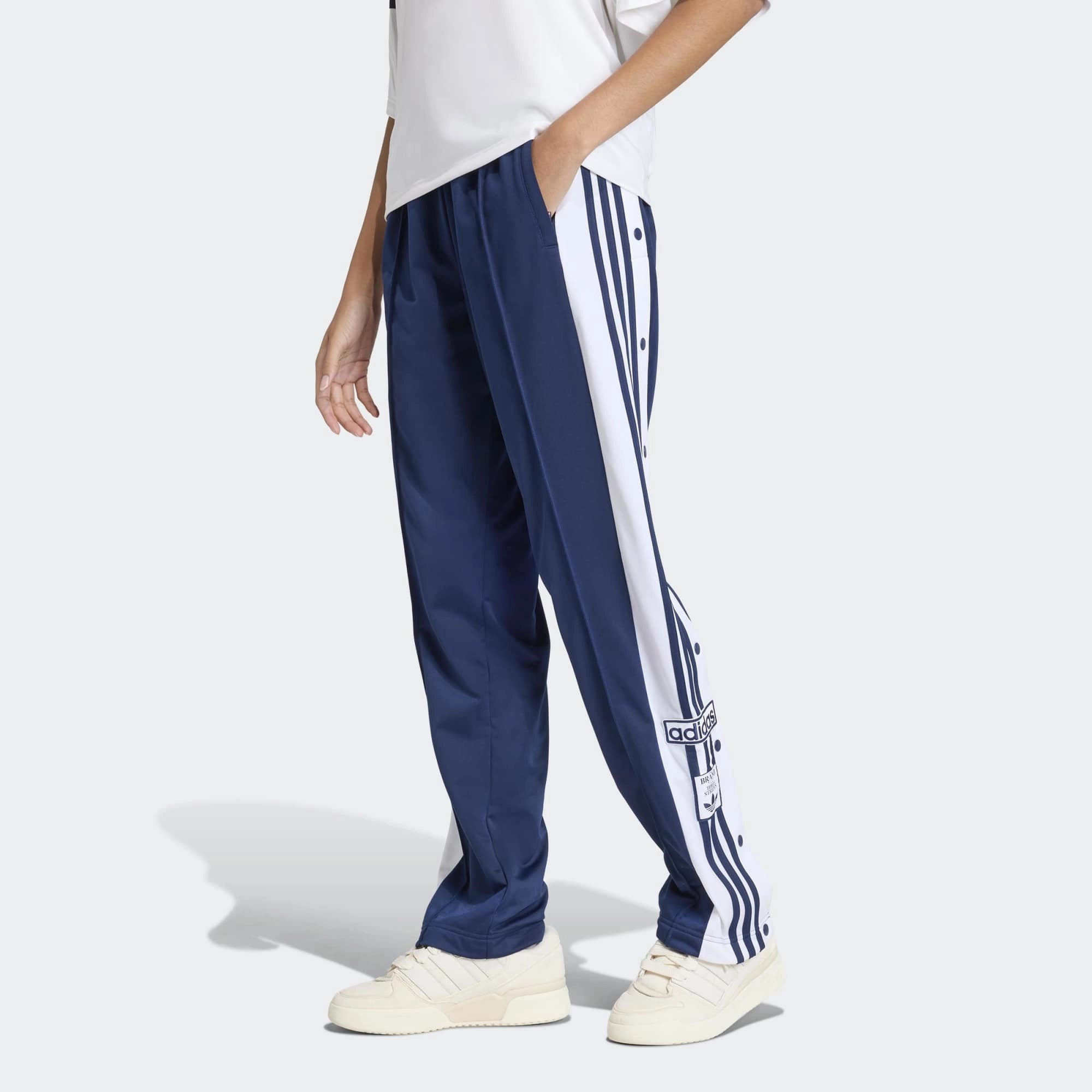 „Dunkelblaue Adidas Originals Adibreak Trainingshose für Damen (Night Indigo) mit durchgehenden Druckknöpfen und weißen Streifen bei Stickabush Berlin.“