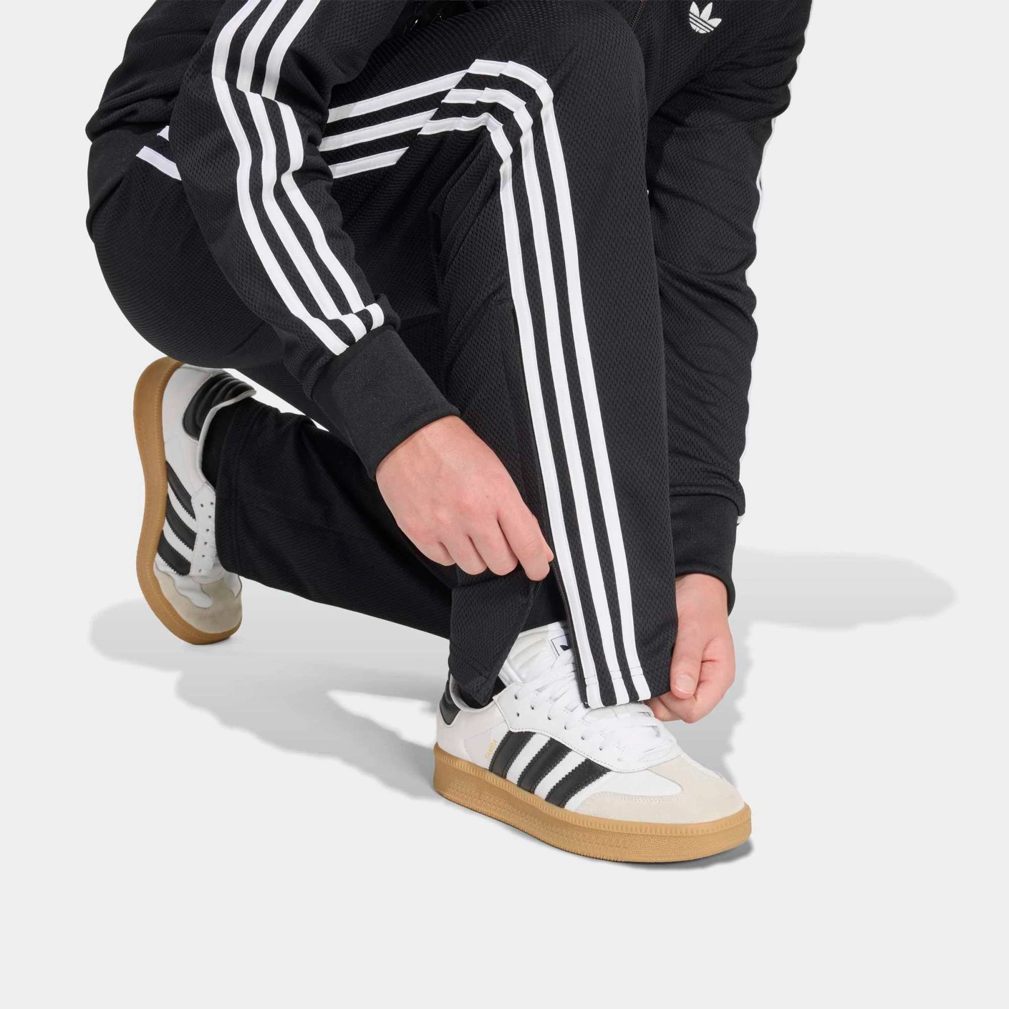 “Adidas Originals Firebird Track Pants Mesh in Schwarz mit weißen Streifen, Frontansicht des Trefoil-Logos und Mesh-Textur bei Stickabush Berlin.“