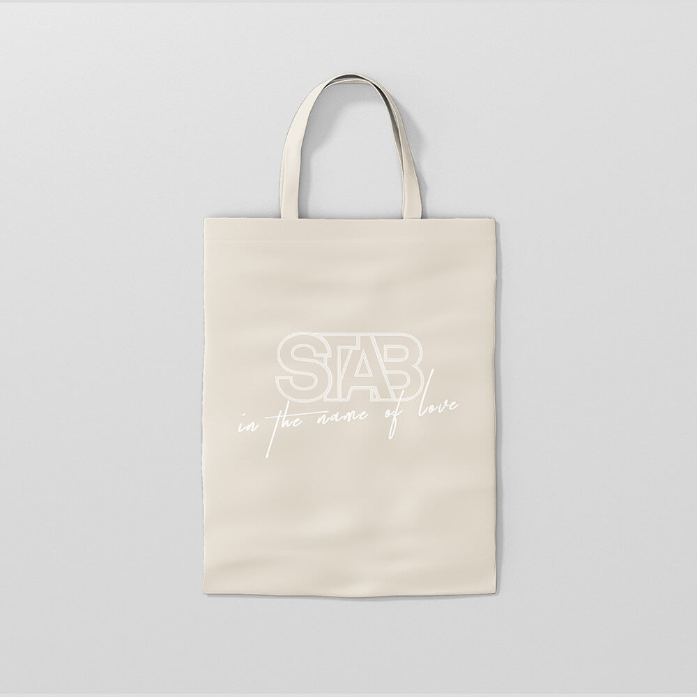 “Cremefarbene STAB "In the Name of Love" Tote Bag aus Stoff mit weißem Schriftzug "In the Name of Love" und Tragegriffen.“
