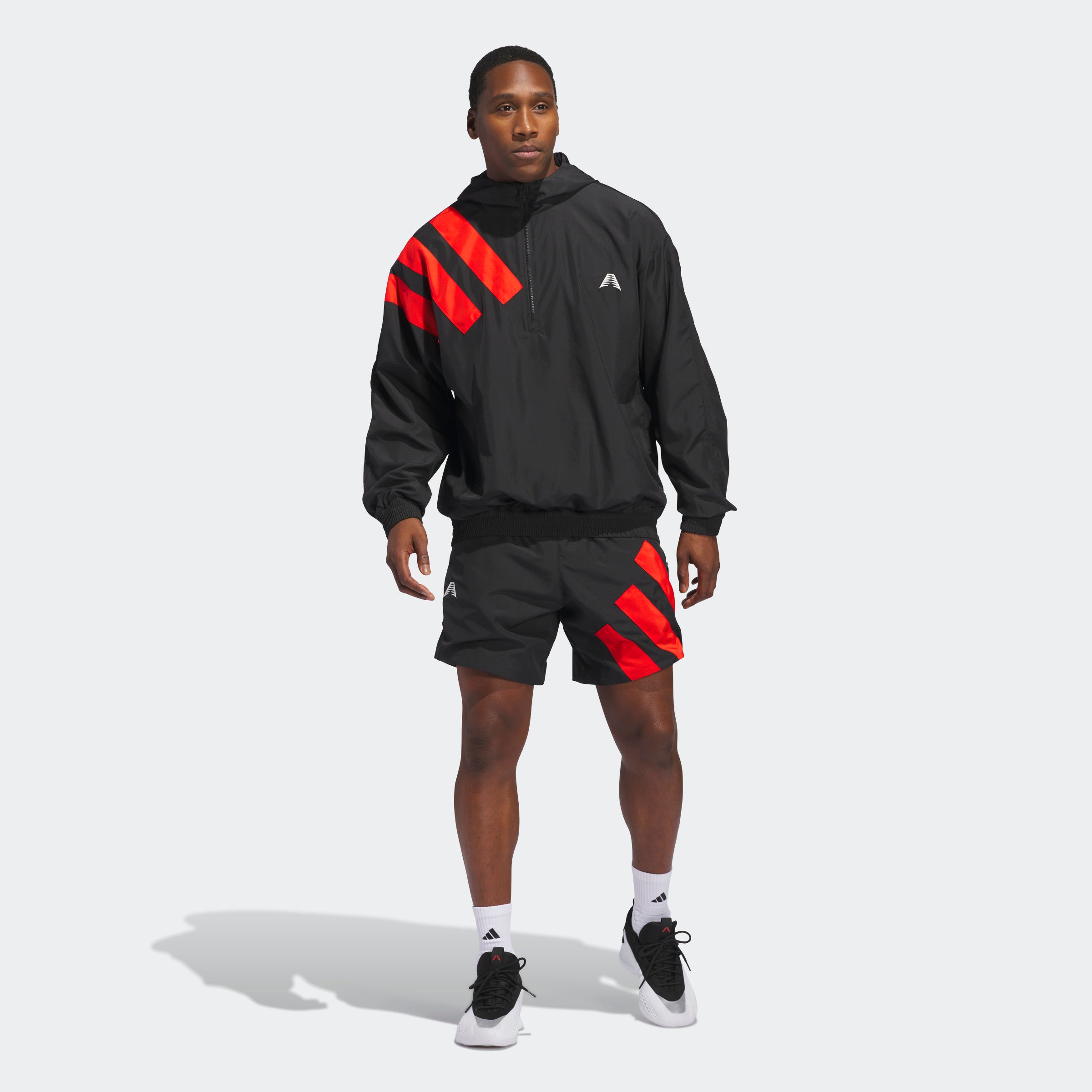 “The Adidas Originals Anthony Edwards Anorak Pullover in black, JC8782, features a hooded design with three bold red diagonal stripes on the upper left shoulder. Made from recycled materials, this durable nylon pullover includes a halfway zipper, white chest logo, and elastic cuffs and waistband for a snug fit.“