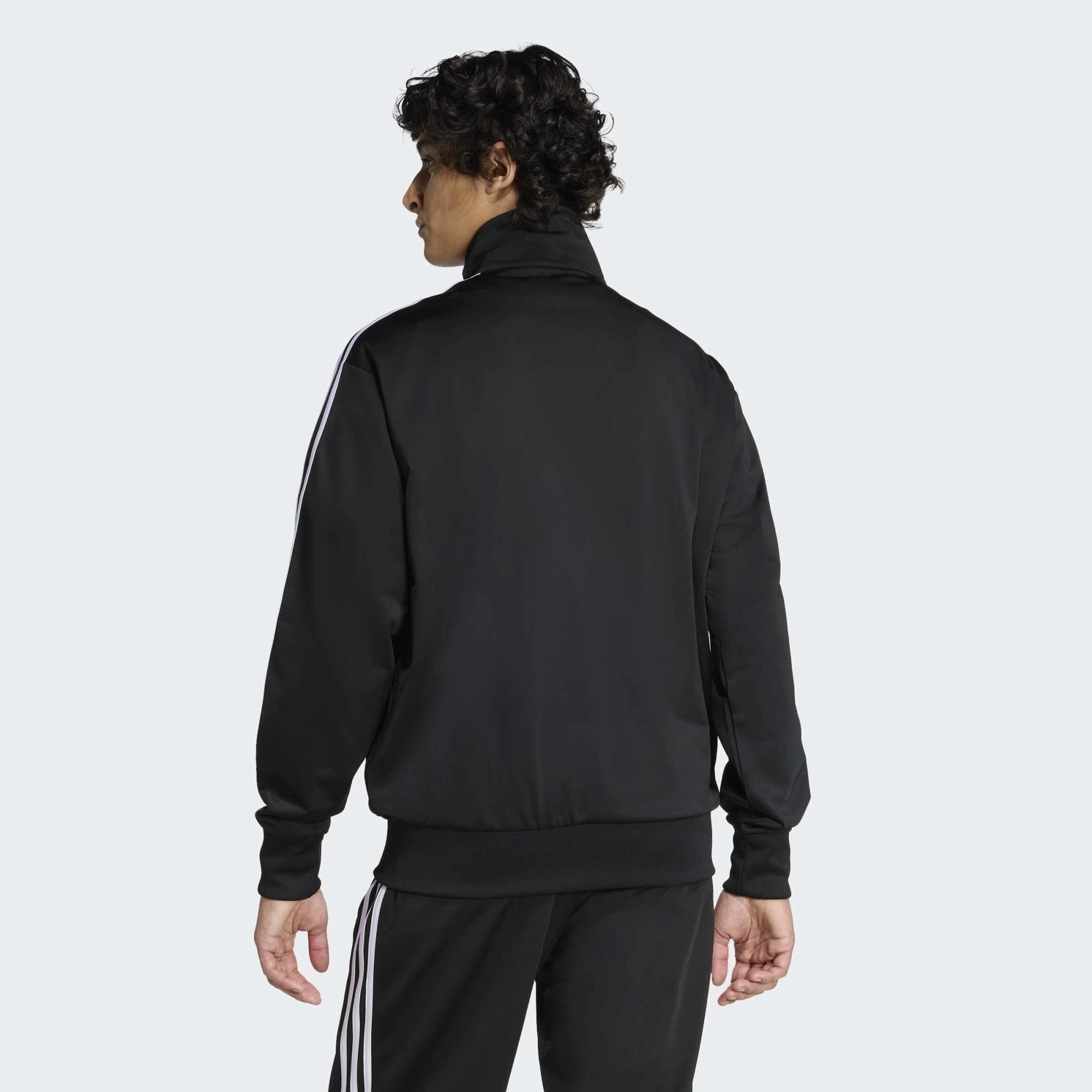 “Klassische adidas Originals Firebird Trainingsjacke in Schwarz mit den charakteristischen drei weißen Streifen an den Ärmeln, Stehkragen und dem gestickten weißen Trefoil-Logo auf der Brust bei Stickabush“.