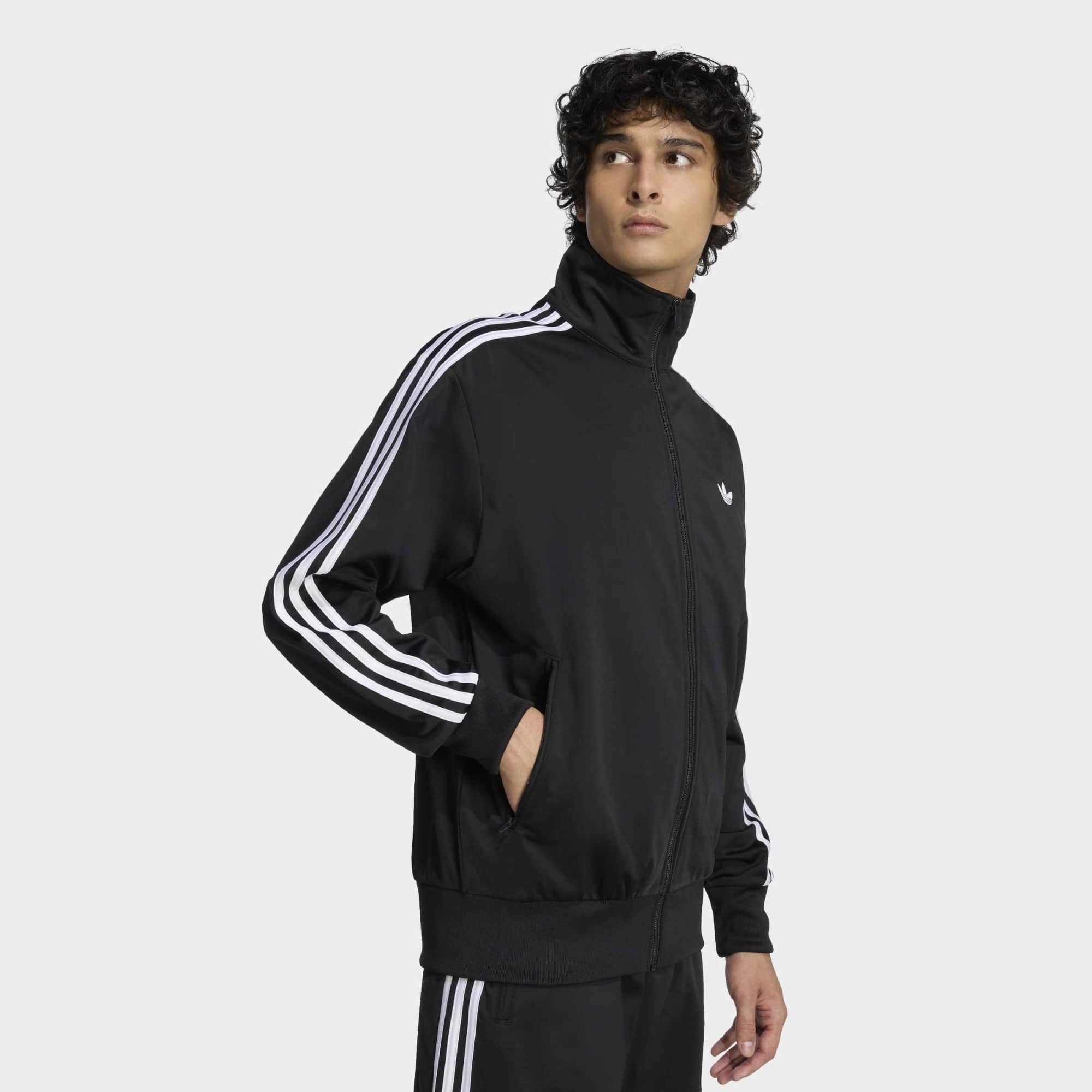 “Klassische adidas Originals Firebird Trainingsjacke in Schwarz mit den charakteristischen drei weißen Streifen an den Ärmeln, Stehkragen und dem gestickten weißen Trefoil-Logo auf der Brust bei Stickabush“.