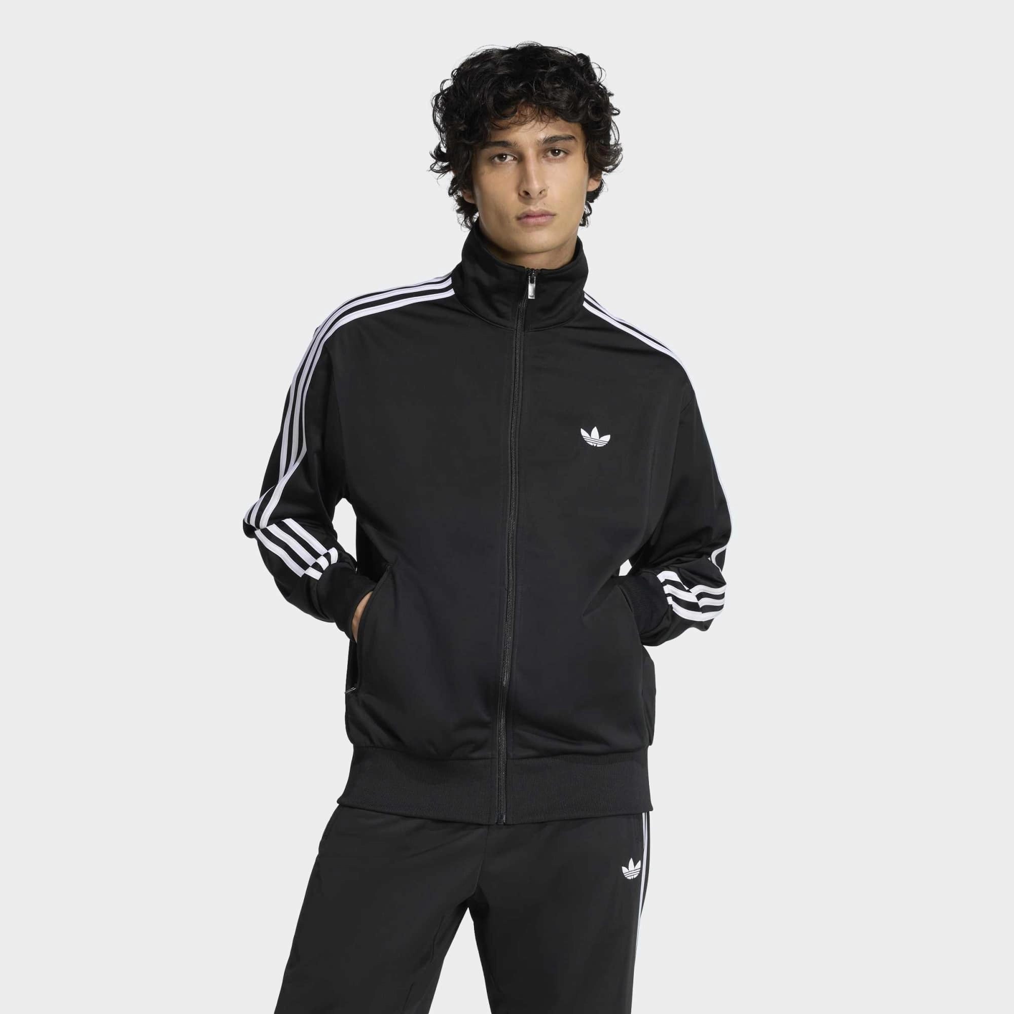 “Klassische adidas Originals Firebird Trainingsjacke in Schwarz mit den charakteristischen drei weißen Streifen an den Ärmeln, Stehkragen und dem gestickten weißen Trefoil-Logo auf der Brust bei Stickabush“.