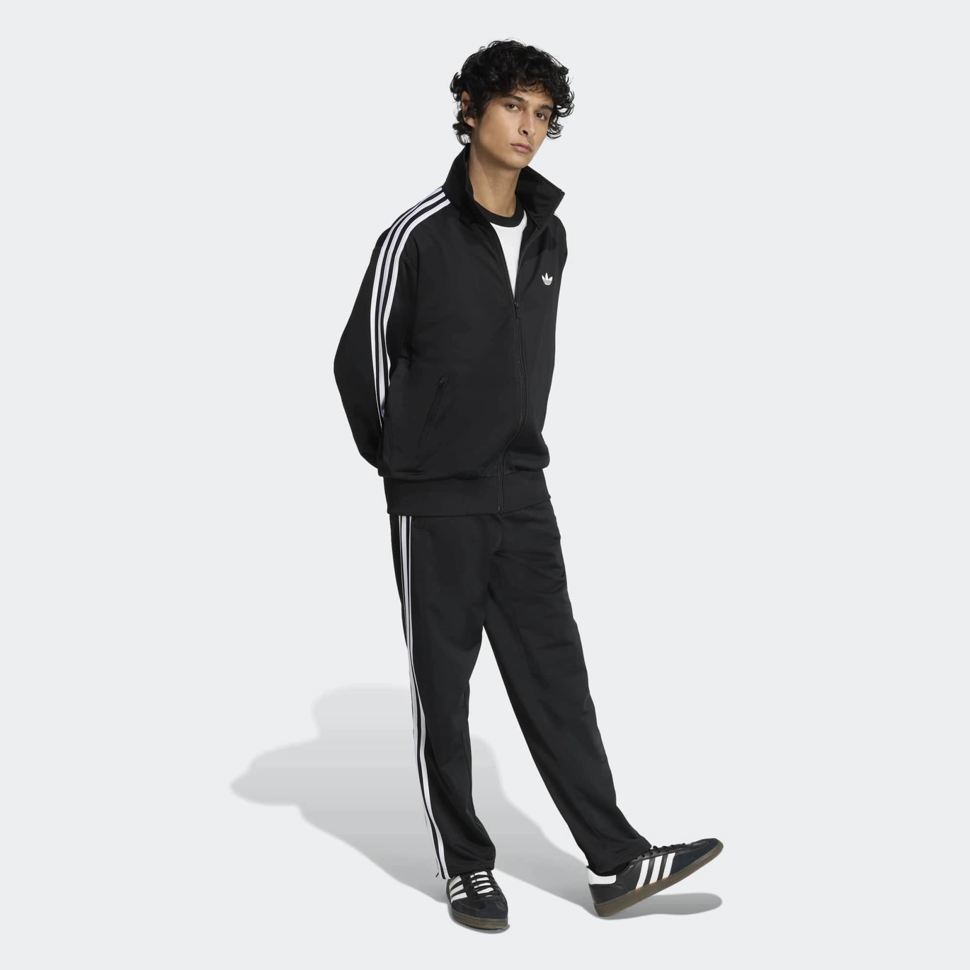 “Klassische adidas Originals Firebird Trainingsjacke in Schwarz mit den charakteristischen drei weißen Streifen an den Ärmeln, Stehkragen und dem gestickten weißen Trefoil-Logo auf der Brust bei Stickabush“.
