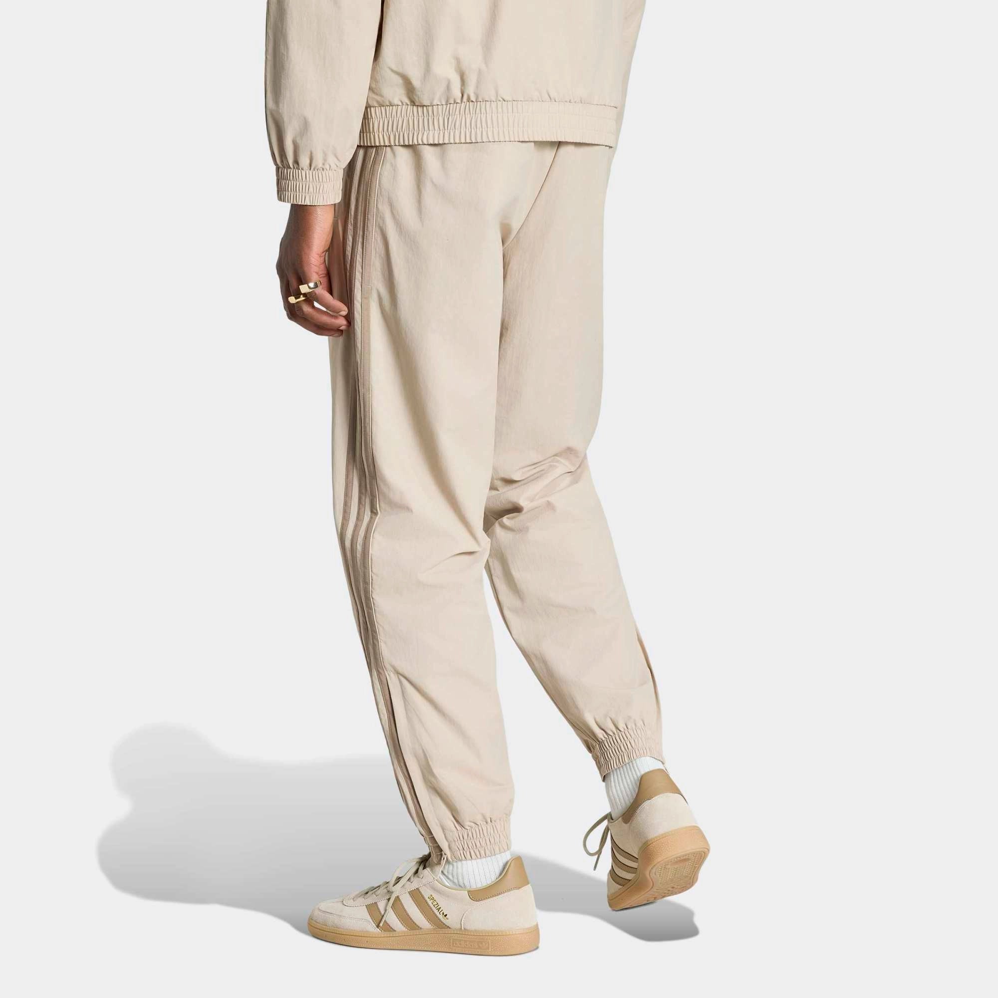 „Beigefarbene adidas Originals Firebird Woven Track Pants (KS7503) aus gewebtem Material mit weißen Streifen – leichte Damen-Trainingshose im Retro-Stil erhältlich bei Stickabush Berlin.“
