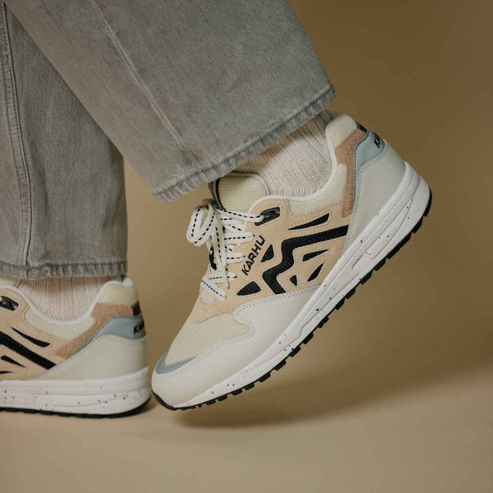 “Die Karhu Legacy 96 - Blanc de Blanc, Sneakers präsentieren sich in einem beigen und hellgrauen Design mit schwarzen Akzenten. Sie verfügen über weiße Schnürsenkel, eine klobige Sohle, einen perforierten Zehenbereich und einen strukturierten Absatz.“