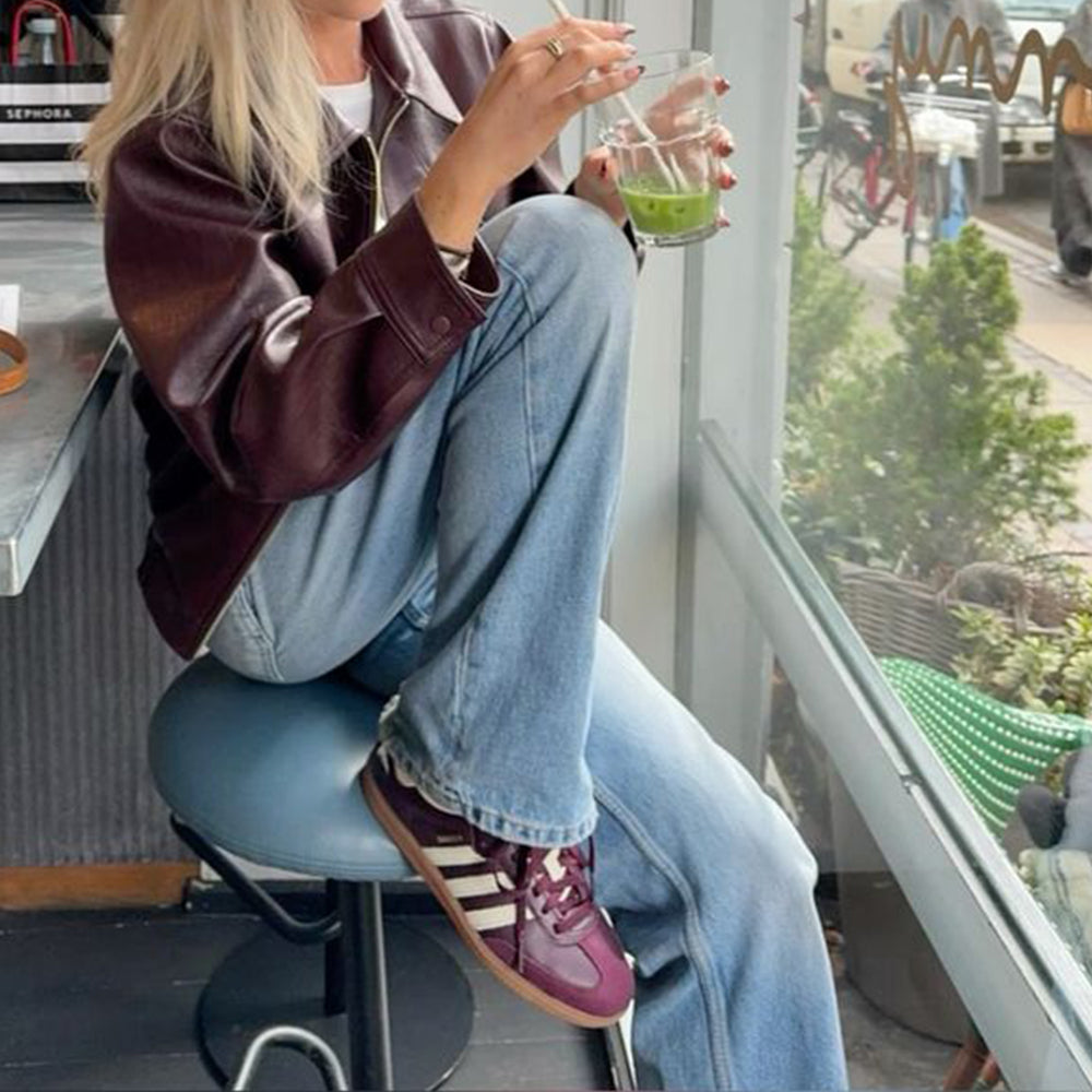 Frau sitzt entspannt in einem Berliner Café mit einem Getränk in der Hand – trägt den adidas Originals Samba OG in Maroon / Cream White (ID0477) von Stickabush Berlin.