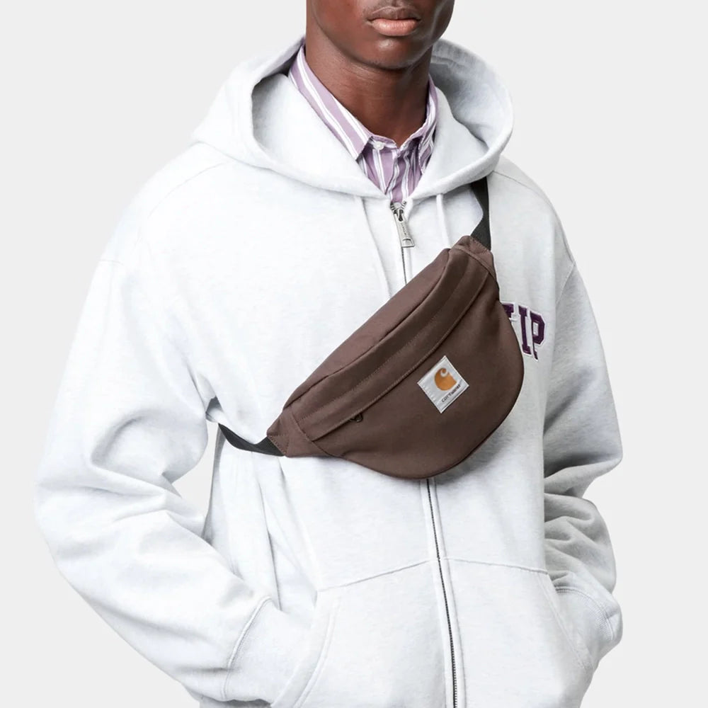 Eine Person trägt eine braune Carhartt WIP Gürteltasche im Crossbody-Stil über einem grauen Zip-Hoodie in einer urbanen Umgebung.