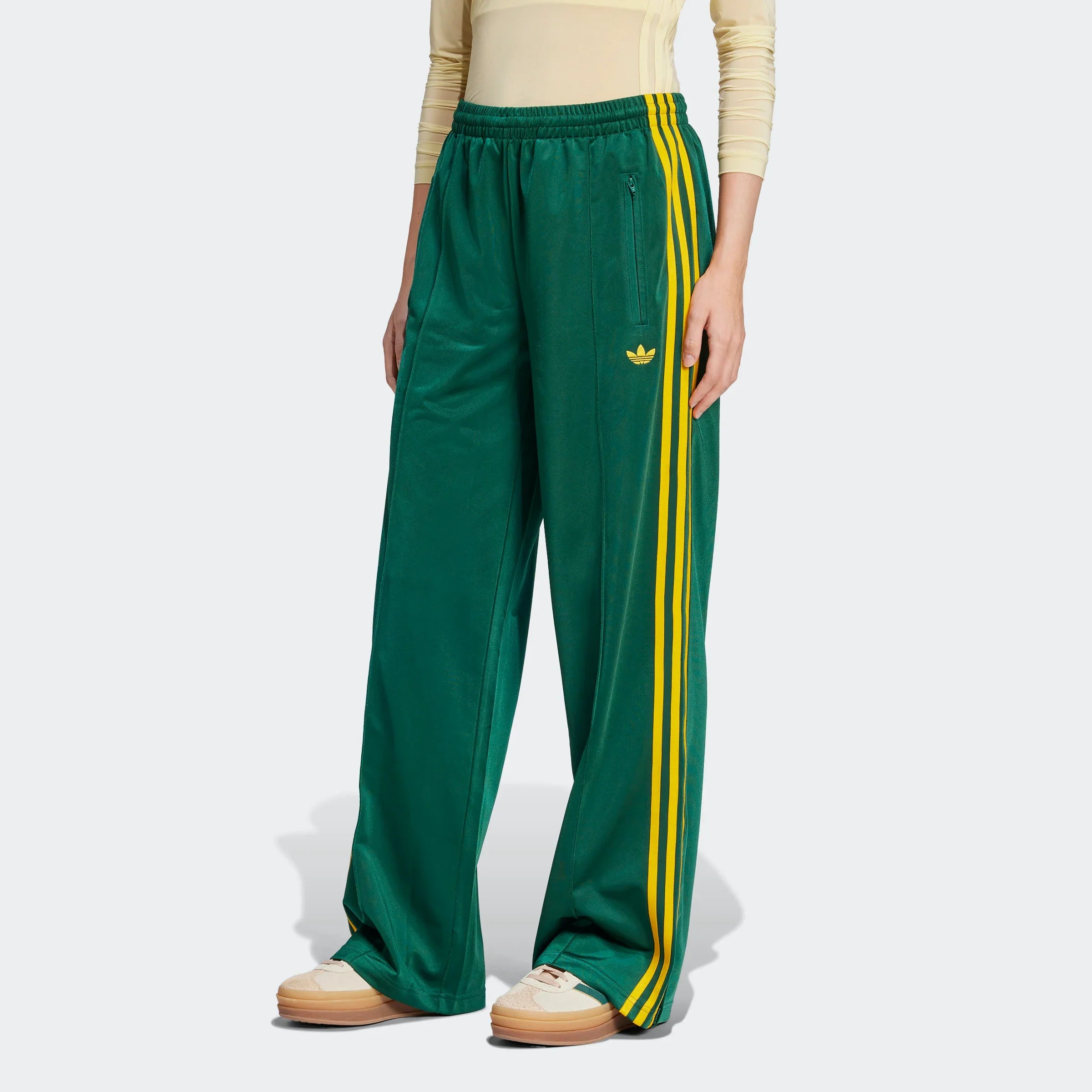 "Grüne Adidas Firebird Trainingshose für Damen mit gelben Streifen. Lockerer Schnitt, glänzendes Trikotmaterial, seitliche Reißverschlusstaschen und kleines Logo."
