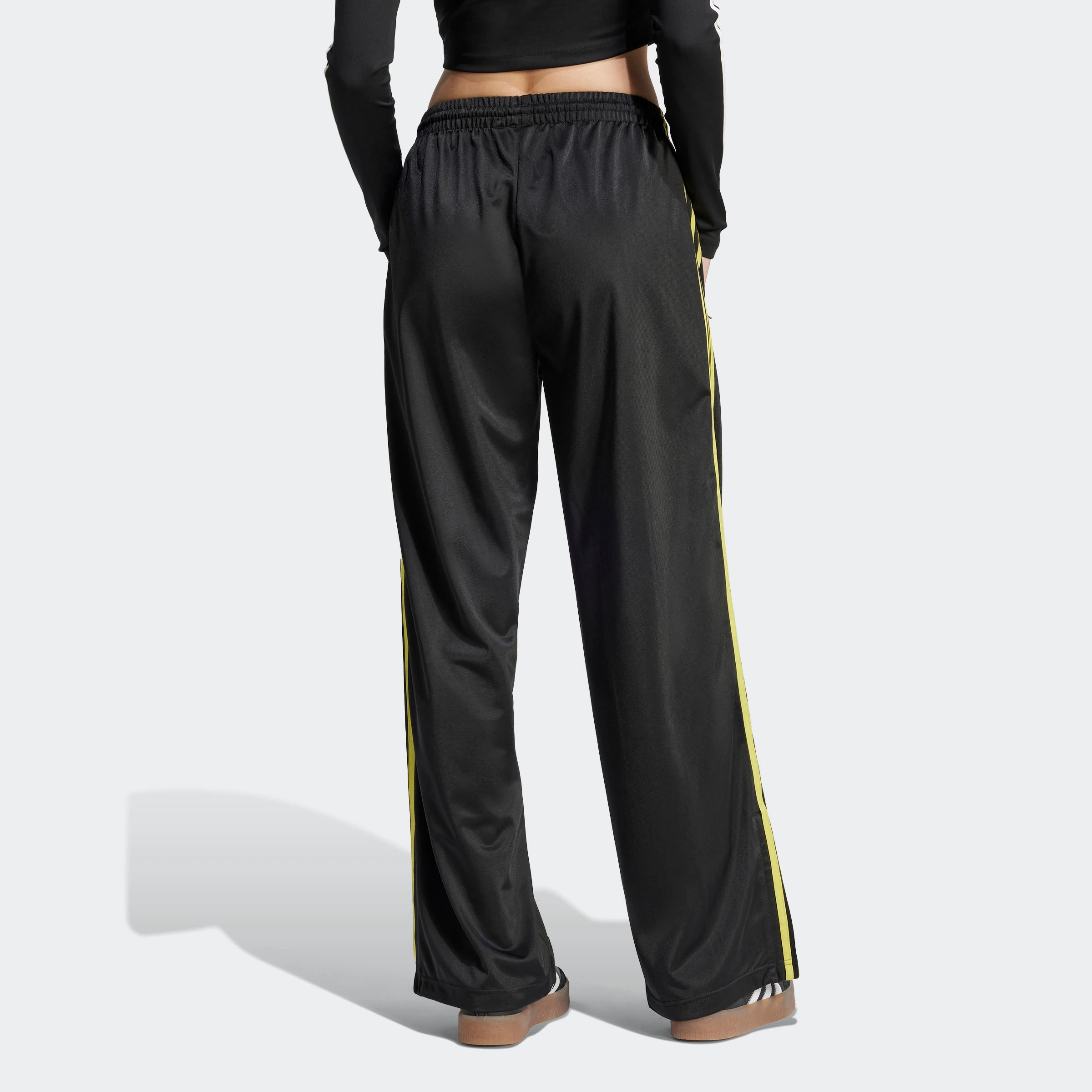 "Schwarze Adidas Firebird Trainingshose für Damen mit hellgelben Seitenstreifen. Lockerer Schnitt, glänzendes Trikotmaterial, seitliche Reißverschlusstaschen, kleines Logo."