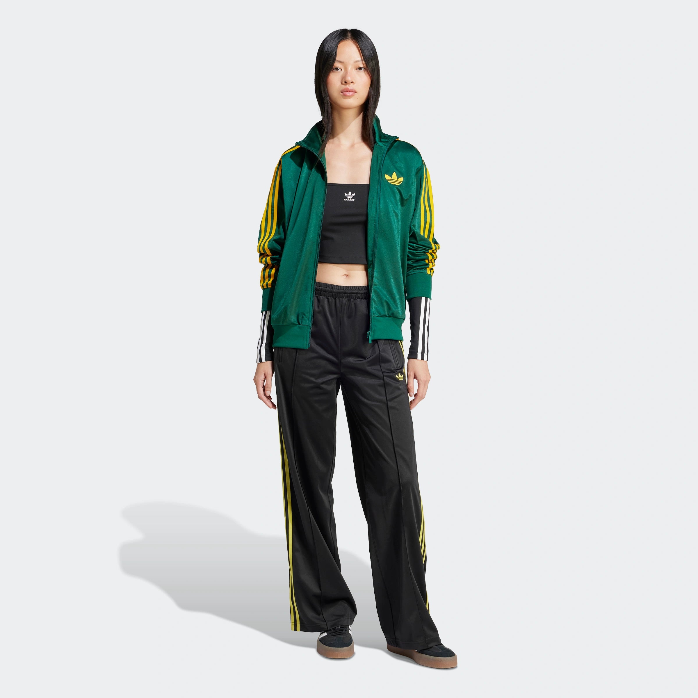 "Schwarze Adidas Firebird Trainingshose für Damen mit hellgelben Seitenstreifen. Lockerer Schnitt, glänzendes Trikotmaterial, seitliche Reißverschlusstaschen, kleines Logo."