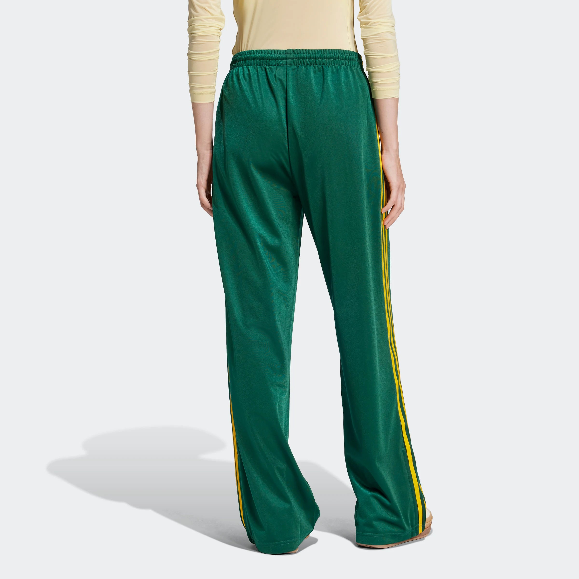 "Grüne Adidas Firebird Trainingshose für Damen mit gelben Streifen. Lockerer Schnitt, glänzendes Trikotmaterial, seitliche Reißverschlusstaschen und kleines Logo."