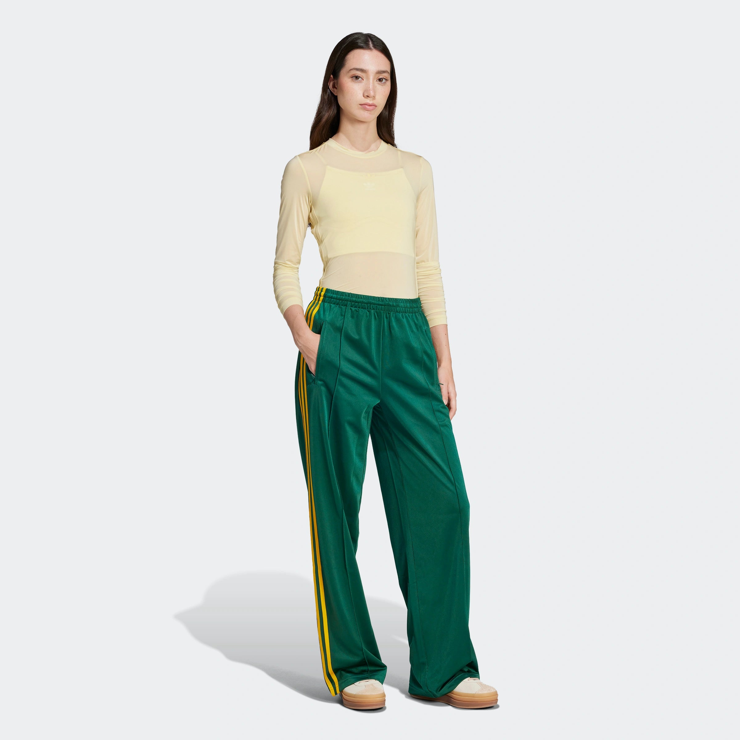 "Grüne Adidas Firebird Trainingshose für Damen mit gelben Streifen. Lockerer Schnitt, glänzendes Trikotmaterial, seitliche Reißverschlusstaschen und kleines Logo."
