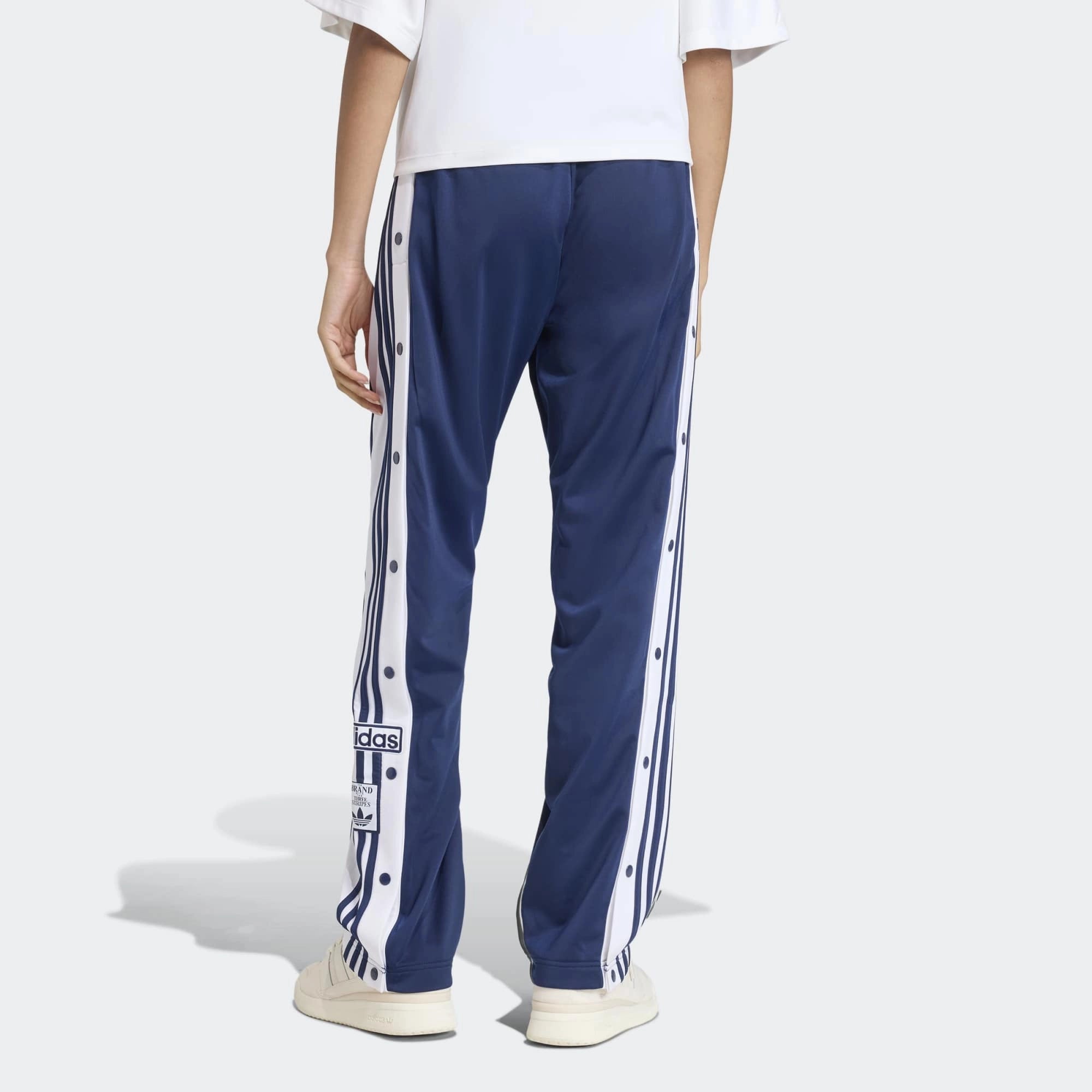 „Dunkelblaue Adidas Originals Adibreak Trainingshose für Damen (Night Indigo) mit durchgehenden Druckknöpfen und weißen Streifen bei Stickabush Berlin.“