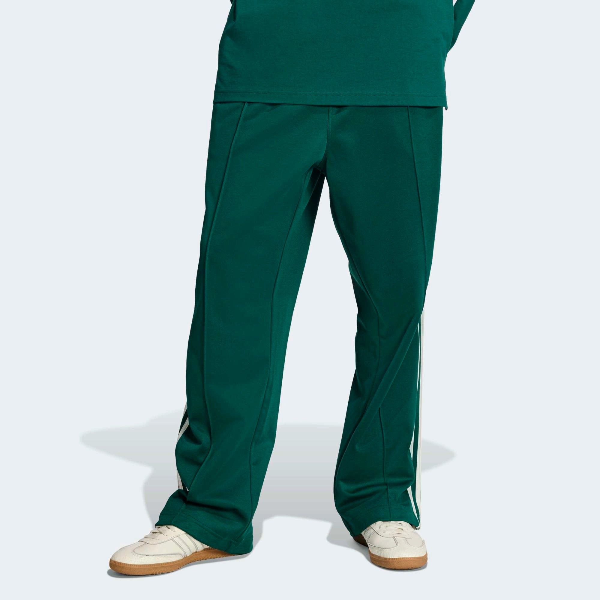 “Vorderansicht Model der adidas Originals Baggy Track Pants in Collegiate Green mit weißen 3-Streifen, seitlichen Reißverschlusstaschen und weitem Beinabschluss bei Stickabush.“