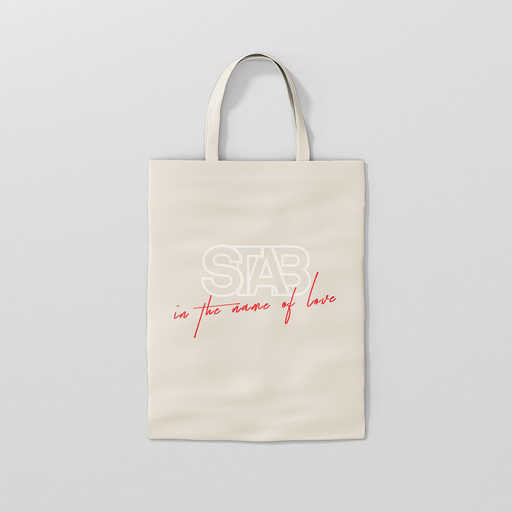 “Cremefarbene STAB "In the Name of Love" Tote Bag aus Stoff mit weißem und rotem Schriftzug "In the Name of Love" und Tragegriffen.“