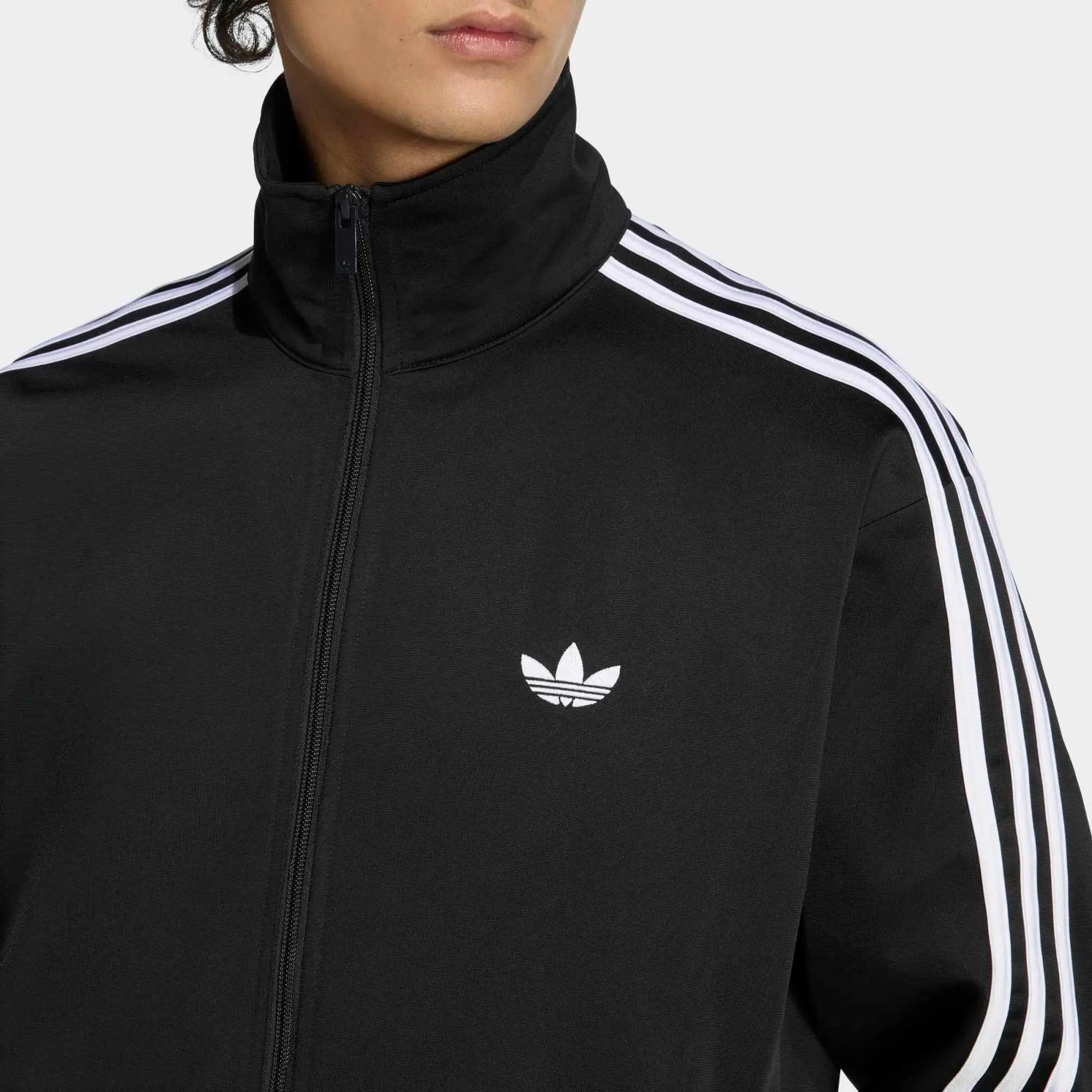 “Klassische adidas Originals Firebird Trainingsjacke in Schwarz mit den charakteristischen drei weißen Streifen an den Ärmeln, Stehkragen und dem gestickten weißen Trefoil-Logo auf der Brust bei Stickabush“.