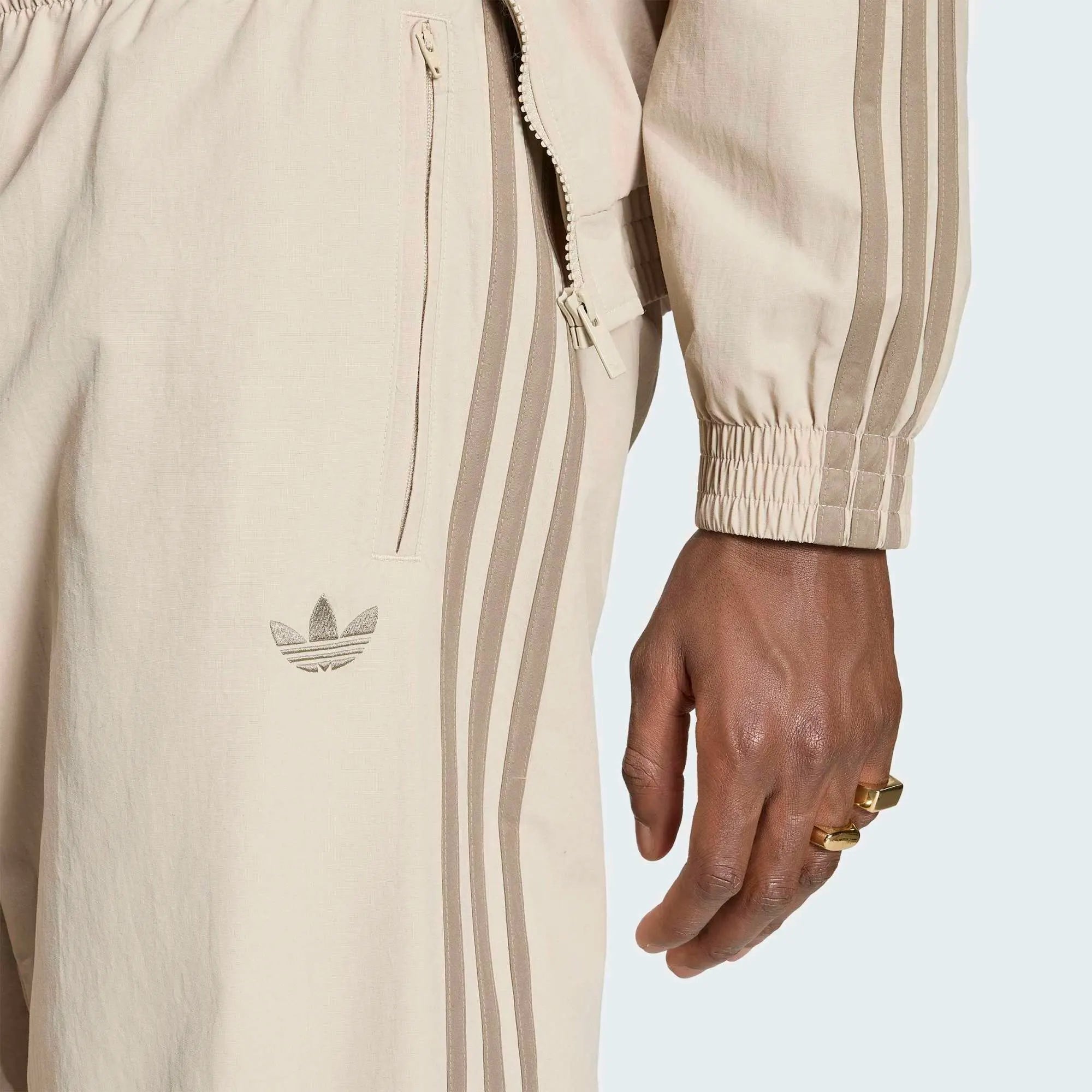 „Beigefarbene adidas Originals Firebird Woven Track Pants (KS7503) aus gewebtem Material mit weißen Streifen – leichte Damen-Trainingshose im Retro-Stil erhältlich bei Stickabush Berlin.“