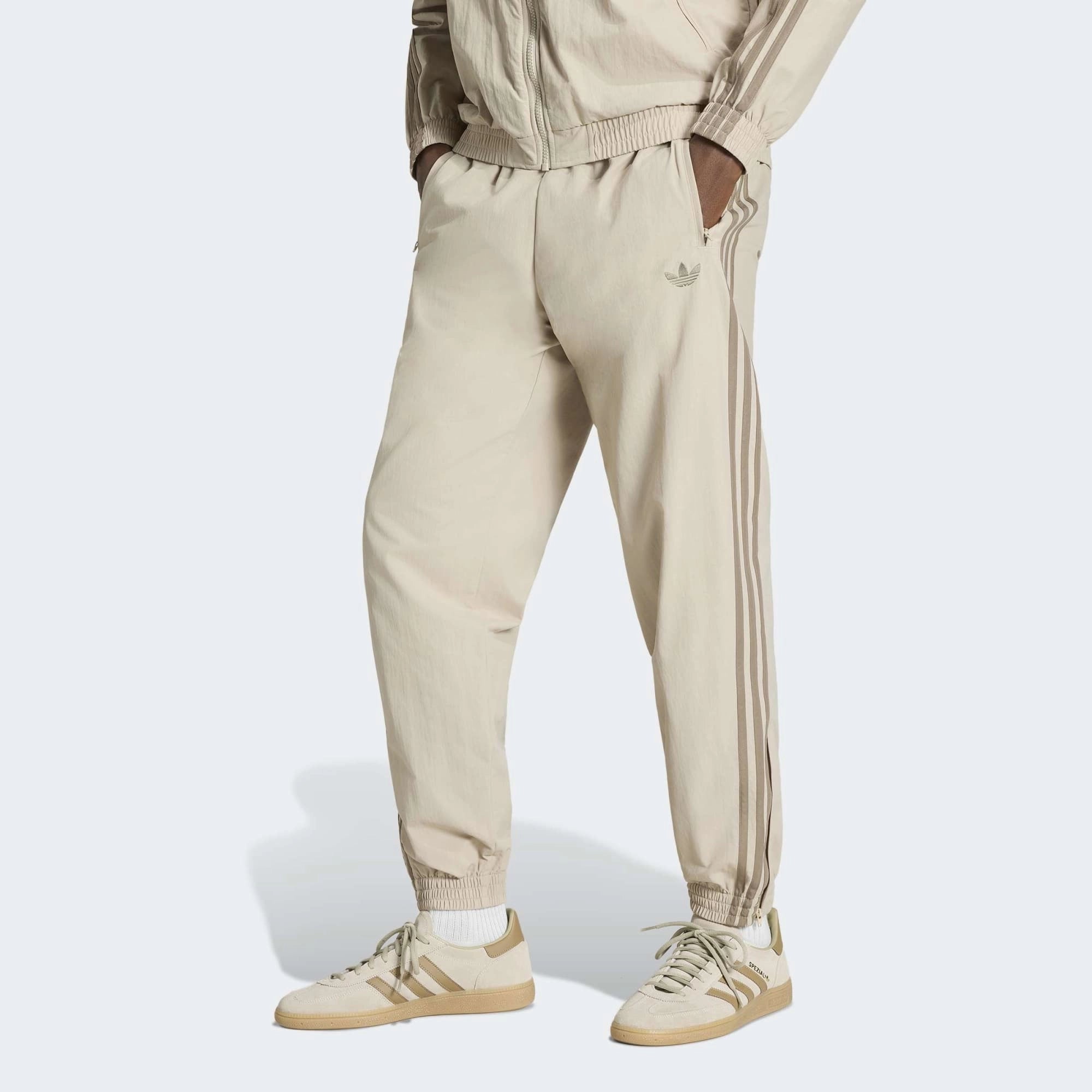 „Beigefarbene adidas Originals Firebird Woven Track Pants (KS7503) aus gewebtem Material mit weißen Streifen – leichte Damen-Trainingshose im Retro-Stil erhältlich bei Stickabush Berlin.“